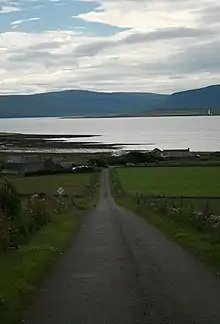 Photographie couleur d’une route descendant en ligne droite vers un bras de mer, avec des collines au fond.