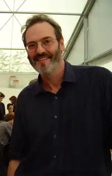 Photo d'un homme en chemise bleu-gris, à la barbe courte et grisonnante, portant des lunettes.