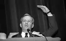 Photographie en noir et blanc d'un homme en costume et faisant un discours devant des micros