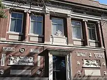 Photographie en couleur de la façade d’un bâtiment. Façade de couleur brique, bas-reliefs blancs de part et d’autre de la porte.