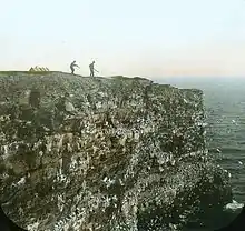 deux hommes avec des fusils sont en haut au bord de la falaise, en train de viser des oiseaux