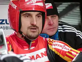 Portrait en buste d'un homme en combinaison rouge, jaune et noir, portant un casque rouge et s'apprêtant à prendre le départ d'une épreuve de luge.