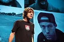 Photo en plan taille de Rodney Mullen, qui se tient devant un écran géant dont l'image affichée est un patchwork de quatre portraits avec un filtre de couleur bleu.