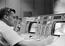 Photographie en noir et blanc d'un homme prenant des notes devant un écran de contrôle.