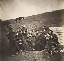 Photographie d'un groupe de soldats prenant la pose devant une cabane semi-enterrée sur le flanc d'une colline. L'un des hommes remplit le verre d'un autre.