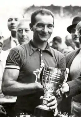 Photographie en noir et blanc d'un cycliste souriant, tenant dans sa main gauche une coupe et donnant une poignée de main à un homme hors champ, la foule entourant ces deux hommes.