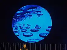 Un homme ridé aux cheveux longs et gris devant un grand écran circulaire sur lequel est diffusé une image d'horloges.