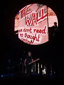 Photographie d'une scène avec un bassiste et un le nom The Wall inscrit derrière lui.