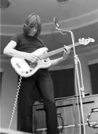 Image monochrome d'un homme jouant de la guitare basse. Il a les cheveux aux épaules, porte une tenue noire et se tient devant un micro.