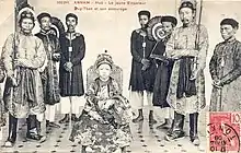Photo ancienne représentant un jeune enfant asiatique, en habit de cour richement décoré, assis sur un trône. Sept homme asiatiques, en habit traditionnel, se tiennent debout à ses côtés.