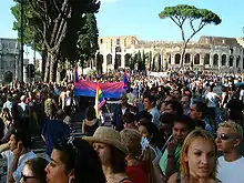 Foule défilant dans la rue, avec les drapeaux arc-en-ciel et bleu/violet/rose au loin