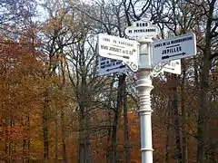 Photographie d'un poteau blanc portant plusieurs panneaux indicatifs de direction, avec des arbres à l'arrière-plan.