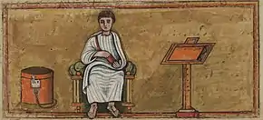 Homme en toge assis, tenant un rouleau de papyrus, un pupitre à sa gauche, une boite à manuscrits à sa droite.