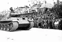 photographie en noir et blanc montrant un char en marche, tandis que le chef de char, émergeant à mi-corps de la tourelle, salue la foule massée au bord de la route.
