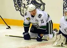 Photographie couleur, un peu floue, d’un hockeyeur en équipement, sur les genous, les points posés sur la glace, tournant la tête vers sa gauche, semblant cligner d’un œil.