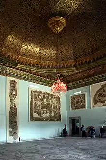 Vue d'une salle d'un palais avec un riche plafond doré et un grand lustre, pièce comportant des mosaïques et des visiteurs à l'arrière-plan