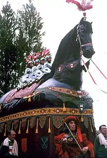 Char de procession représentant un immense cheval noir harnaché et décoré, sur lequel sont positionnés quatre hommes en amure.