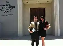 Man with light skin and dark hair and woman with dark hair and light skin stand in front of a building with a sign that reads The Church of Jesus Christ of Latter-day Saints; Visitors Welcome