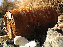 Photo en couleurs d’une canette de boisson rouillée et percée, abandonnée sur un terrain caillouteux.