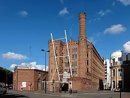 Photographie en couleur représentant un bâtiment industriel en brique rouge de cinq étages vu en enfilade, flanqué d'une tour crénelée à gauche et d'une tour plus étroite prolongée en cheminée, elle aussi crénelée, à droite
