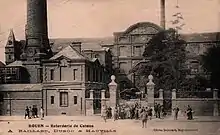 Carte postale noir et blanc. Façades de plusieurs bâtiments appartenant à une même usine. Grande cheminée tronquée par la prise de vue. Devant le vaste portail d'entrée, un groupe probablement d'employés.