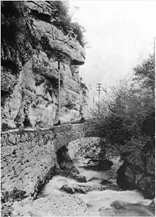 Une route à flancs de montagne se poursuit par un pont qui franchit un torrent.