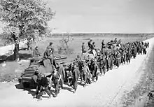 Sur une route de campagne, des soldats dans des véhicules militaires observent le passage d'un groupe d'hommes à pied.