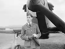 Photographie en noir et blanc du buste d'un homme en uniforme, portant une casquette et des gants, debout devant un avion de chasse