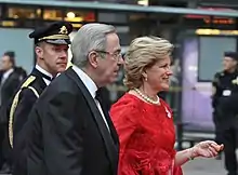 Photographie en couleurs d'un homme et d'une femme assez âgés en tenue de soirée. Derrière eux se tient un garde en uniforme.