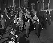 Photographie noir et blanc de deux hommes quittant une salle suivi par de nombreuses personnes.