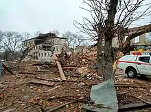 Photo des ruines d'un bâtiment de trois étages éventré par une bombe, de nombreux débris jonchant le sol, et des arbres nus cassés en morceaux.