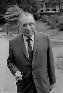 photographie d'un homme dans un parc paysager, il porte des lunettes, un costume et une cravate. Dans sa main droite un porte-cigarette.