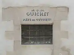 Fenêtre étroite percée dans un mur, sur lequel est inscrit « M.O.M. Guichet Paye des ouvriers ».