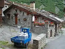 La photo couleur montre un petit triporteur motorisé bleu chargé de 4 bidons de lait en aluminium. La scène est située dans un village aux maisons de pierre et aux toits en dalle de pierre et ardoises.