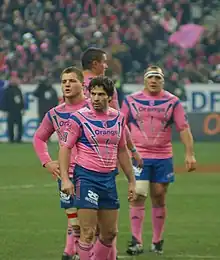 Photographie en couleurs. Quatre joueurs portant un maillot rose et bleu sont debout, avec Christophe Dominici en premier plan, regardant vers la gauche les sourcils froncés.