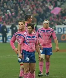 Au premier plan quatre joueurs sont statiques lors d'un temps mort, vêtus de rose et de bleu. Au fond, on devine les spectateurs parisiens avec les mêmes couleurs en tribune.