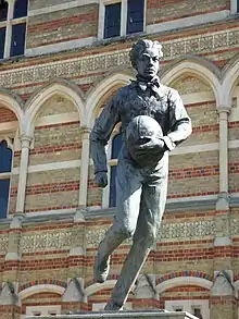 Photographie en couleurs montrant une statue en bronze représentant un homme courant ballon sous le bras, devant les murs des bâtiments de l'école.