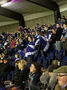 Gros plan dans une tribune sur des supporters écossais attentifs à la rencontre en cours. Au premier plan, les spectateurs sont assis sagement, dans des tenues classiques. Au second, la population, plus jeune, est debout, quelques-uns ont du maquillage, d'autres des drapeaux.