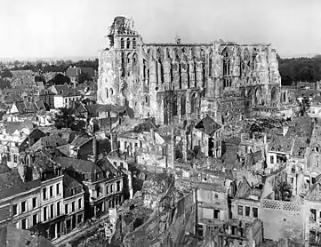 Photographie en noir et blanc. Basilique avec sa charpente et ses parties supérieures détruites. Au premier plan, nombreuses maisons en ruines.