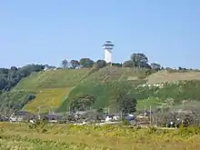 Photo couleur d'une colline couvetre de verdure, avec un village à son pied, et une tour blanche à son sommet, sur fond de ciel bleu.