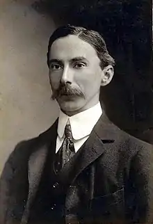 Photographie en noir et blanc d'un homme au regard fixe, portant une moustache en col blanc et cravate, semblant figé dans une attitude rigide.