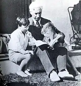 Photographie en noir et blanc d'un homme aux cheveux blancs assis, un livre ouvert sur les genoux et deux enfants à ses côtés : une fillette au large sourire à sa gauche, penchée en avant et à sa droite un petit garçon aux cheveux bruns, pieds nus ayant l'index gauche dans la bouche.