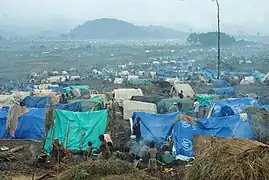 Un camp de réfugiés aux nombreuses tentes, certaines portant les couleurs des Nations Unies. En arrière plan des montagnes zaïroises.