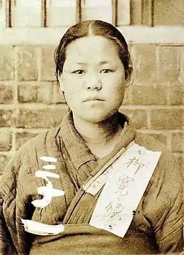 Photographie d'une très jeune femme faisant face au photographe. Elle porte un vêtement de toile grossier, et sur la clavicule d'un morceau de tissu est accroché, avec son nom écrit en kanji.