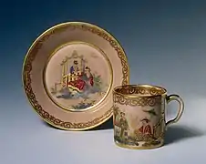Une tasse et une sous-tasse en porcelaine blanche et liseré doré, représentant une scène de famille.