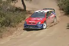 Photo de la Citroën Xsara WRC de Sébastien Loeb, à la livrée rouge, blanche et bleue, arrivant de face en dérapage dans une virage droite.