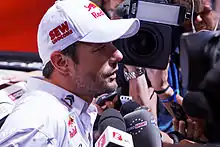 Sébastien Loeb de profil avec une casquette, entouré par les micros de plusieurs journalistes.