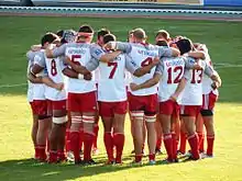 Des joueurs de rugby habillés de t-shirt se regroupent en cercle, se tenant par les épaules.