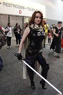 Femme rousse habillée en noir et tenant une réplique de sabre laser