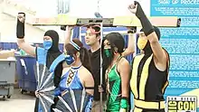 Cinq personnes dont 4 déguisées en personnages du jeu posent pour une photo pendant une convention. La personne centrale est habillée en noir et porte une grande barre sur la tête, qui représente l'interface de jeu de Mortal Kombat